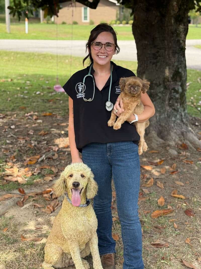 A woman wearing a black shirt and stethoscope stands outdoors, holding a small brown dog and standing beside a larger cream-colored dog.