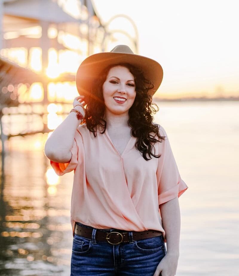 Woman with curly dark hair wearing a hat and light pink blouse stands by water at sunset, smiling, with a structure and sunlight in the background.