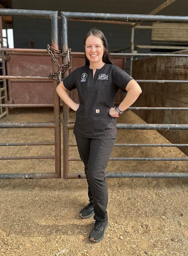 A person in black scrubs stands smiling with hands on hips in front of metal livestock gates in a barn setting.