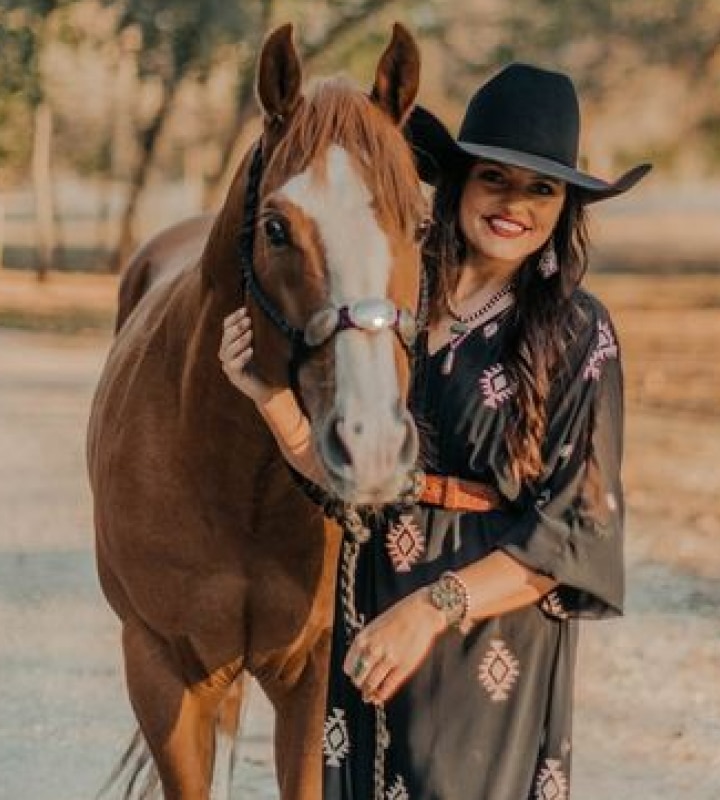 Woman in a black hat and patterned dress stands next to a brown horse outdoors.