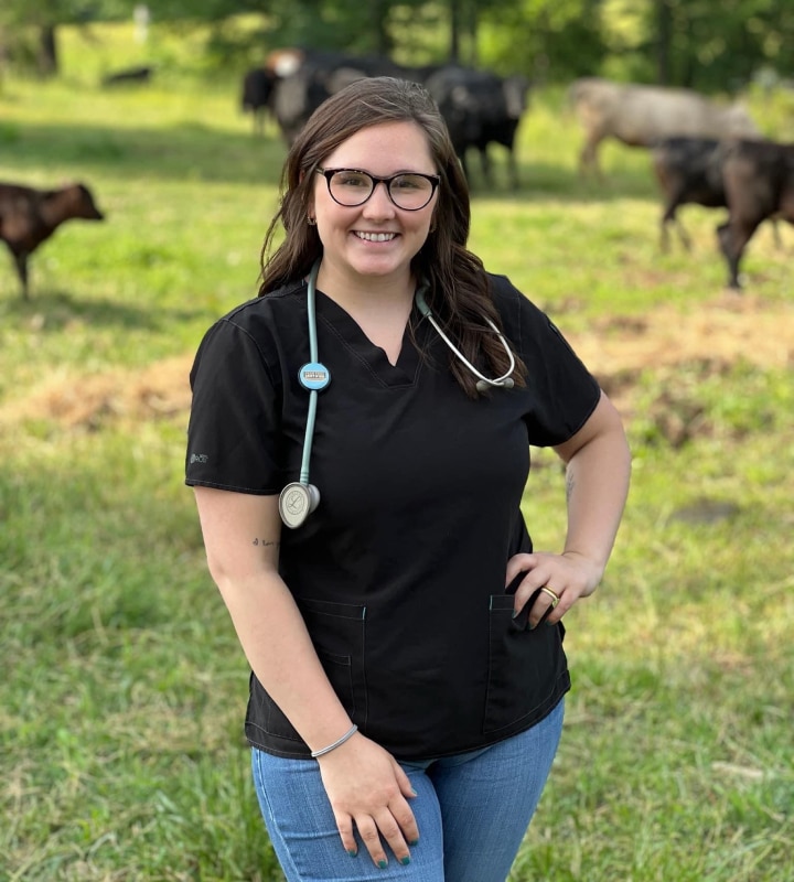 A person in a black uniform with a stethoscope stands in a grassy field with cows in the background.