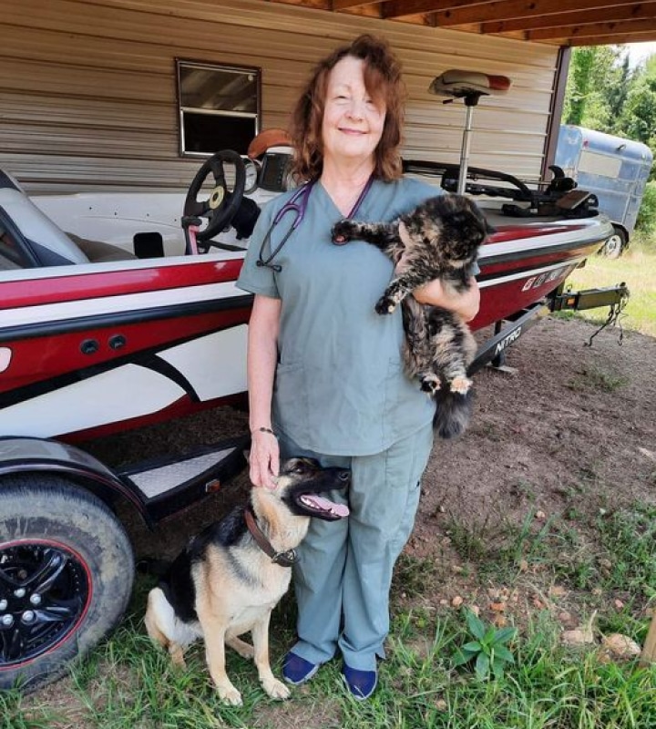 A woman in scrubs stands outside holding a cat, with a dog sitting by her side and a fishing boat in the background.