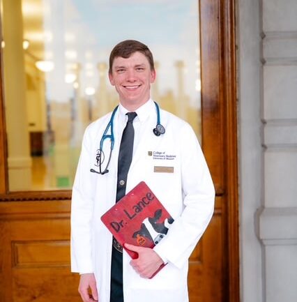 A man in a white lab coat and stethoscope stands outside a building, holding a red folder labeled "Dr. Lance" with a cartoon image of a cow.