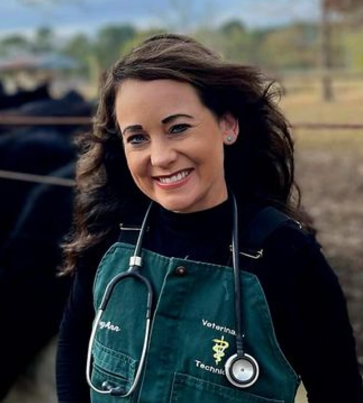 A smiling woman in a green veterinary technician uniform stands outdoors, wearing a stethoscope around her neck.